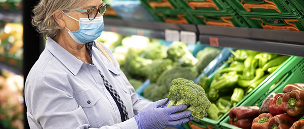 Senior woman wearing face mask and rubber gloves selects broccoli and other vegetables  in a supermarket -active elderly pensioners with trolley