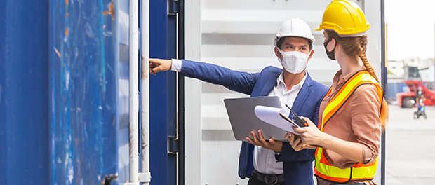 Teamwork foreman man and woman wearing protection face mask and safety helmet using laptop and holding clipboard checking containers in cargo ship for import export, Industrial container cargo concept.